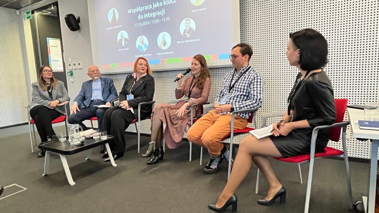 Panel dyskusyjny pod nazwą "Współpraca jako klucz do integracji". Cztery kobiety i dwóch mężczyzn siedzi na czerwonych krzesłach, w tle grafika promująca panel. Na grafice zdjęcia profilowe panelistów. Jedna z kobiet mówi przez mikrofon. Wydarzenie odbywa się w Plus Jeden.