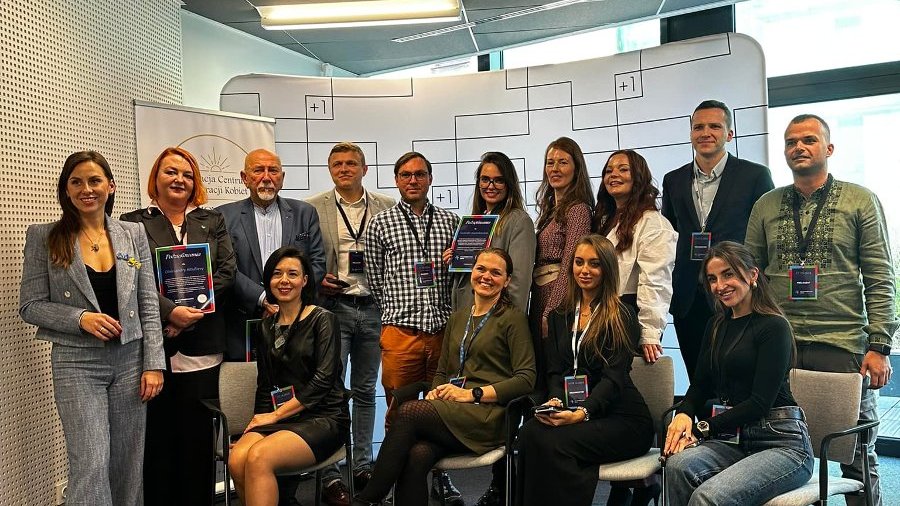 Pozowane zdjęcie grupowe organizatorów i prelegentów dnia ukraińskiego podczas Dni Przedsiębiorczości Poznań 2024. Pięciu mężczyzn i dziewięć kobiet stoi i siedzi czekając na zrobienie zdjęcia. W tle baner promujący przestrzeń pracy wspólnej Plus Jeden.