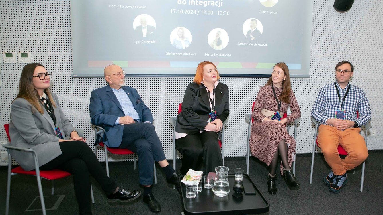 Panel dyskusyjny pod nazwą "Współpraca jako klucz do integracji". Trzy kobiety i dwóch mężczyzn siedzi na czerwonych krzesłach, w tle grafika promująca panel. Na grafice zdjęcia profilowe panelistów. Wydarzenie odbywa się w Plus Jeden.