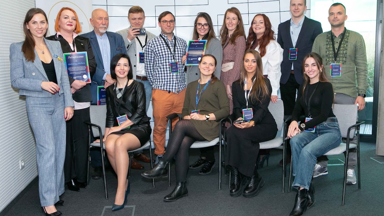 Organizatorzy i prelegenci konferencji pod nazwą "Twój czas na zmianę: od inspiracji do działania" pozują do grupowego zdjęcia. Dziewięć kobiet i pięciu mężczyzn. Uśmiechając się stoją i siedzą na krzesłach czekając na zrobienie zdjęcia. W tle baner promujący przestrzeć pracy wspólnej Plus Jeden.