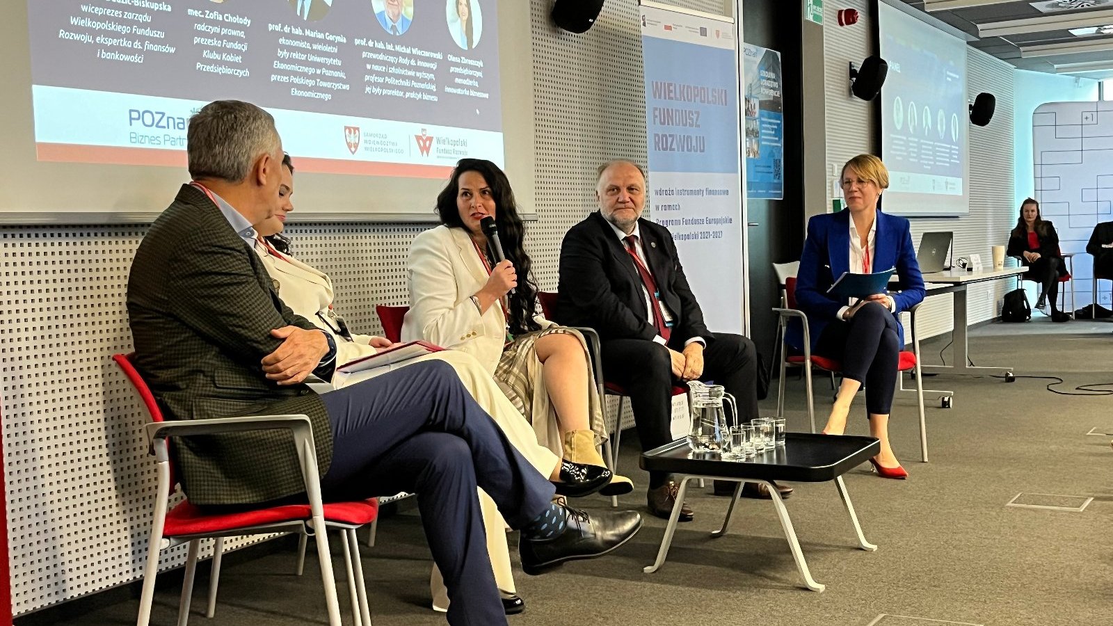 Panel dyskusyjny podczas konferencji "Finanse dla biznesu". Trzy kobiety i dwóch mężczyzn siedzi na czerwonych krzesłach. Jedna kobieta mówi przez mikrofon. Przed nimi czarny stolik z dzbankiem z wodoą oraz szklankami. W tle wyświetlana grafika dotycząca panelu dyskusyjnego przedstawiająca zdjęcia profilowe panelistów oraz roll-up promujący Wielkopolski Fundusz Rozwoju.