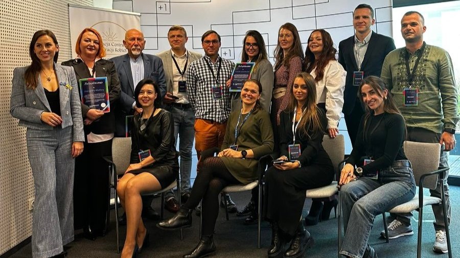 Pozowane zdjęcie grupowe organizatorów i prelegentów dnia ukraińskiego podczas Dni Przedsiębiorczości Poznań 2024. Pięciu mężczyzn i dziewięć kobiet stoi i siedzi czekając na zrobienie zdjęcia. W tle baner promujący przestrzeń pracy wspólnej Plus Jeden.