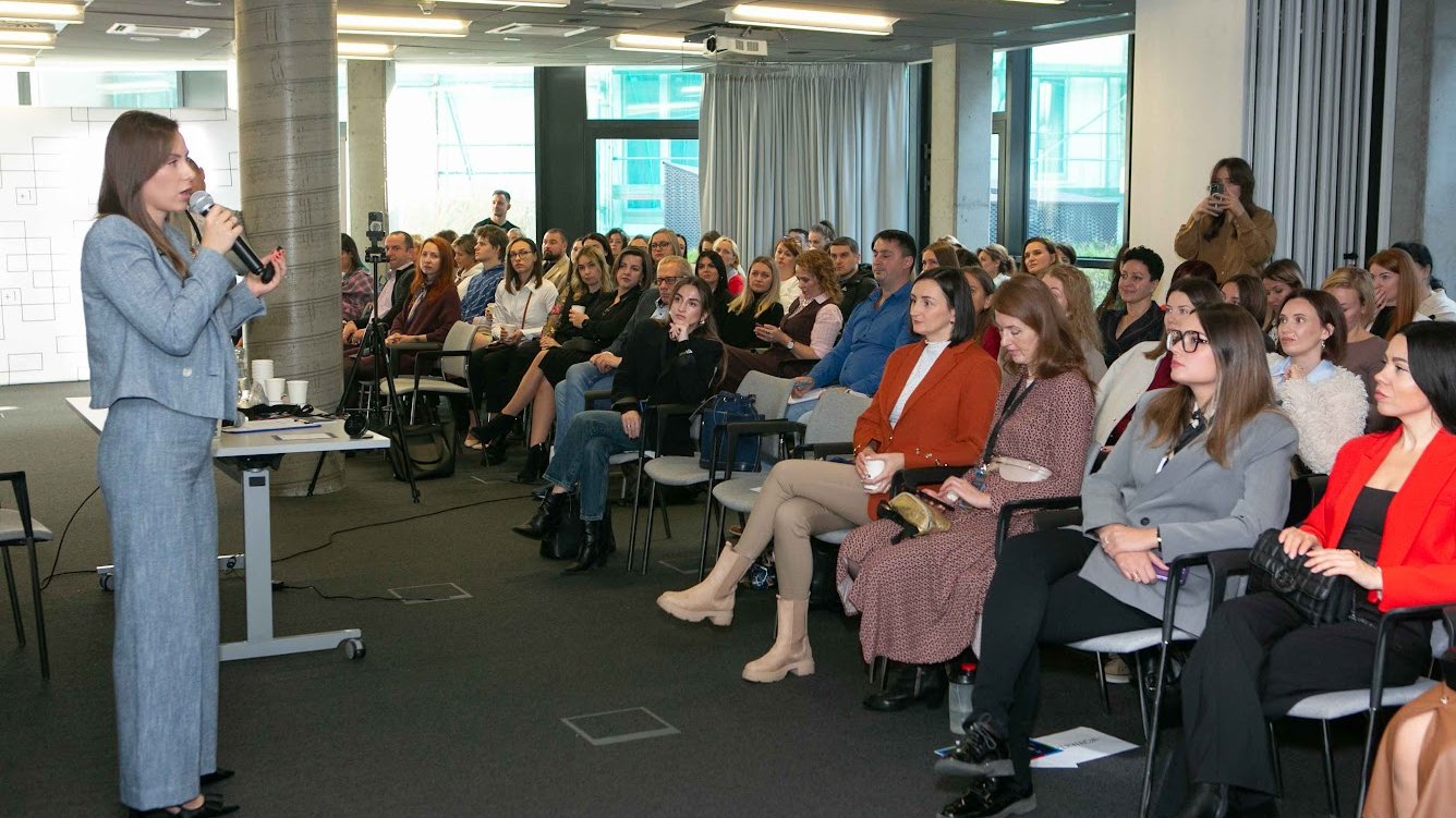 Jedna z prelegentek przemawia do audytorium w podczas konferencji dla obywateli Ukrainy. Widać dużo ludzi siedzących na krzesłach. Jedna osoba w tle robi zdjęcie prowadzącej.