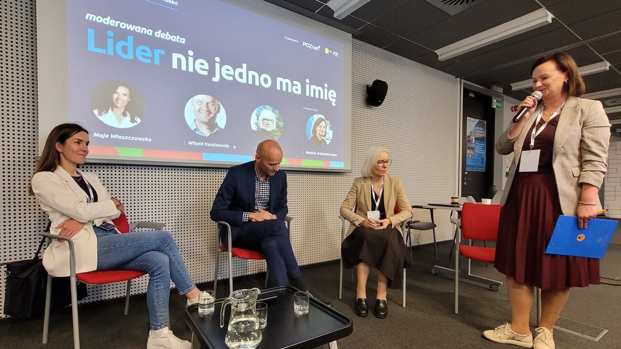 Panel dyskusyjny podczas konferencji "Lider na horyzoncie". Dwie kobiety i jeden mężczyzna siedzą na czerwonych krzesłach, podczas, gdy moderatorka panelu stoi i ich przedstawia. Pierwsza od lewej to Maja Włoszczowska. Przed nimi stolik z wodą a w tle wyświetlana jest grafika promująca panel dyskusyjny.