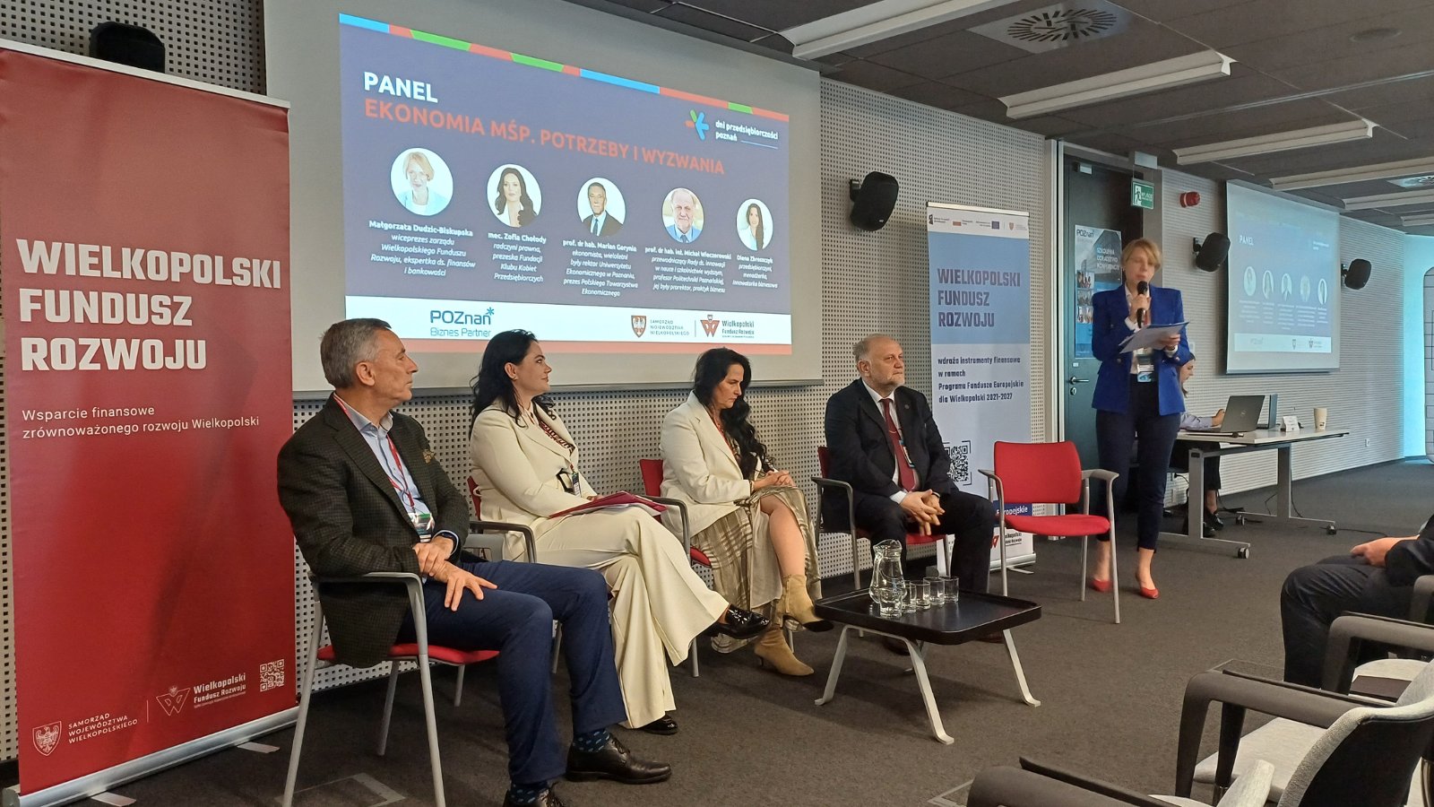 Panel dyskusyjny podczas konferencji "Finanse dla biznesu". Dwie kobiety i dwóch mężczyzn siedzą na czerwonych krzesłach. Jedna kobieta stoi i mówi przez mikrofon. Przed nimi czarny stolik z dzbankiem z wodą oraz szklankami. W tle wyświetlana grafika dotycząca panelu dyskusyjnego przedstawiająca zdjęcia profilowe panelistów oraz roll-up promujący Wielkopolski Fundusz Rozwoju.