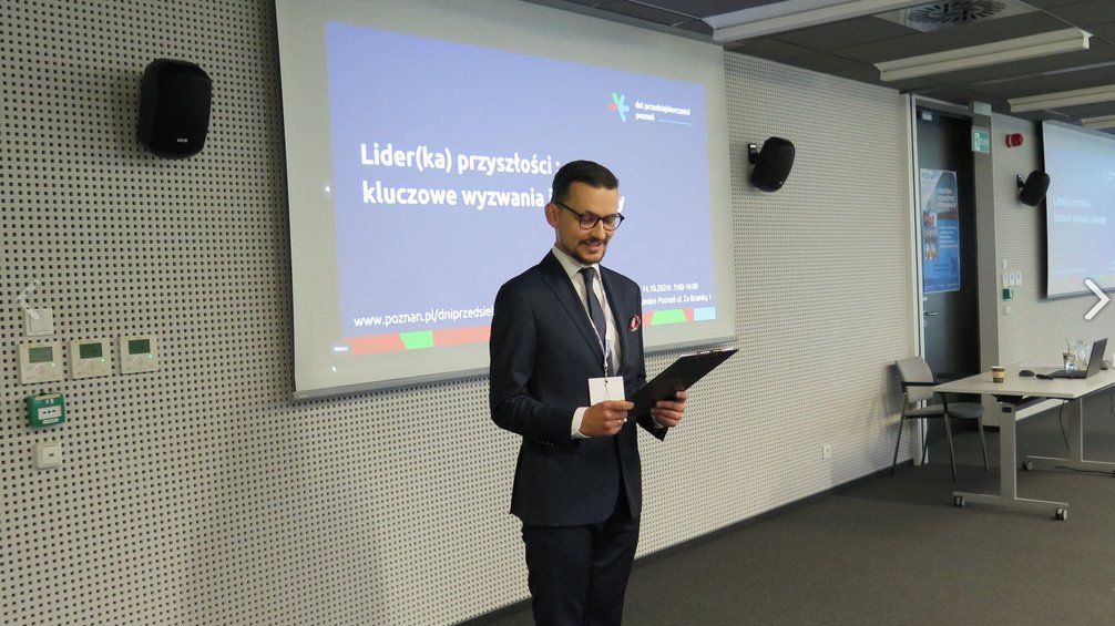 Konferansjer zapowiada, czytając z kartki kolejnego prelegenta podczas konferencji "Lider (ka) przyszłości: kluczowe wyzwania i dylematy". W tle wyświetlana grafika promująca konferencję. W oddali biurko i krzesło.
