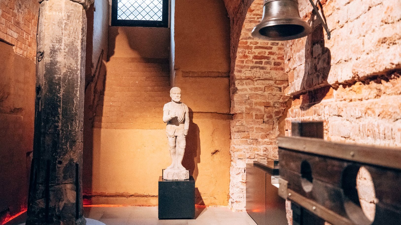 Figura rycerza, obok cokół, na którym stała rzeźba. Po prawej stronie, na ceglanym murze zawieszony średniej wielkości dzwon. - grafika artykułu