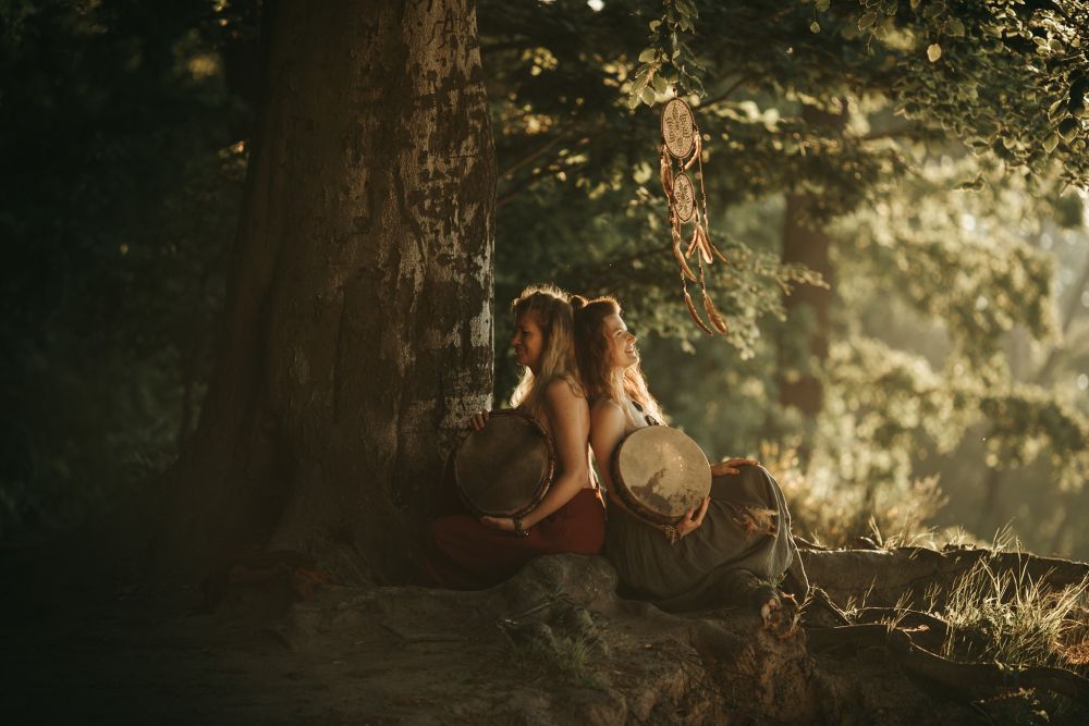 Dwie dziewczyny w sukienkach siedzą pod dużym drzewem, opierają się o siebie plecami, każda z nich trzyma w ramionach staroświecki bęben. Jedną z nich oświetla słońce. - grafika artykułu