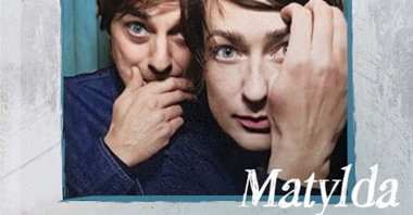 Zdjęcie kobiety i mężczyzny w niebieskiej ramce, wokół zdjęcia szare, szerokie obramowanie, u dołu zdjęcia napis "Matylda. Łukasiewicz". Na zdjęciu kobieta dotyka dłonią krótkich włosów, a mężczyzna za nią dotyka dłoniąswoich ust.