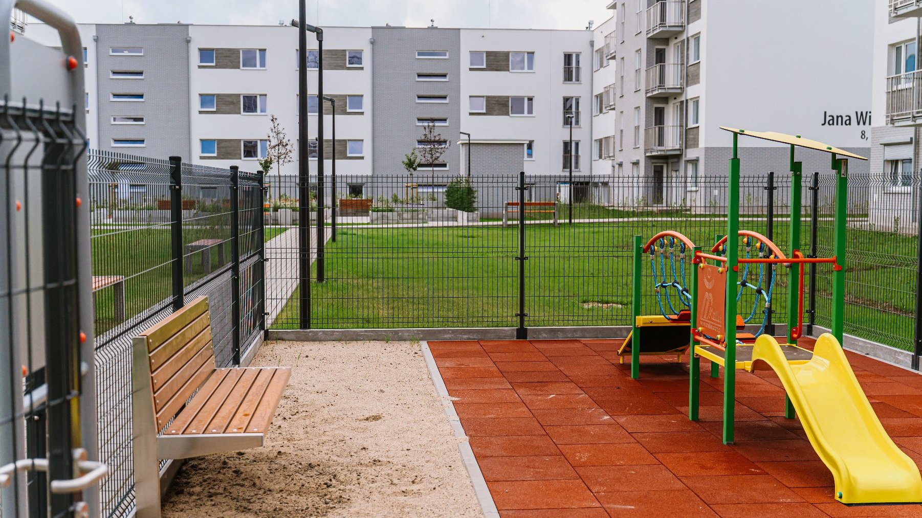 Na zdjęciu widać plac zabaw. Na placu znajduje się ślizgawka dla małych dzieci i ławka. Plac jest ogrodzony. W tle zdjęcia widać budynki mieszkalne i trawnik.