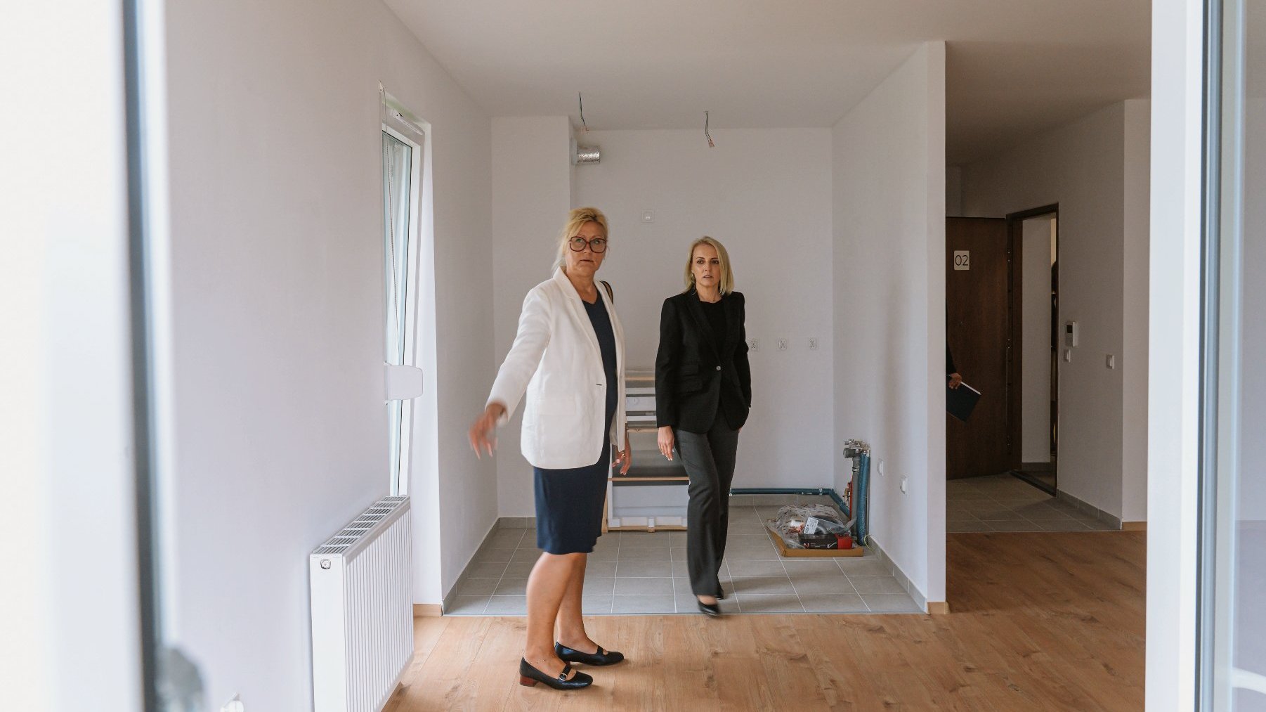 Na zdjęciu widać wnętrze mieszkania. Ściany w mieszkaniu są białe. Część podłogi jest wyłożona brązowymi panelami, a część kafelkami. Drzwi wejściowe do mieszkania są otwarte. Jest na nich numer 02. Mieszkanie oglądają dwie kobiety.