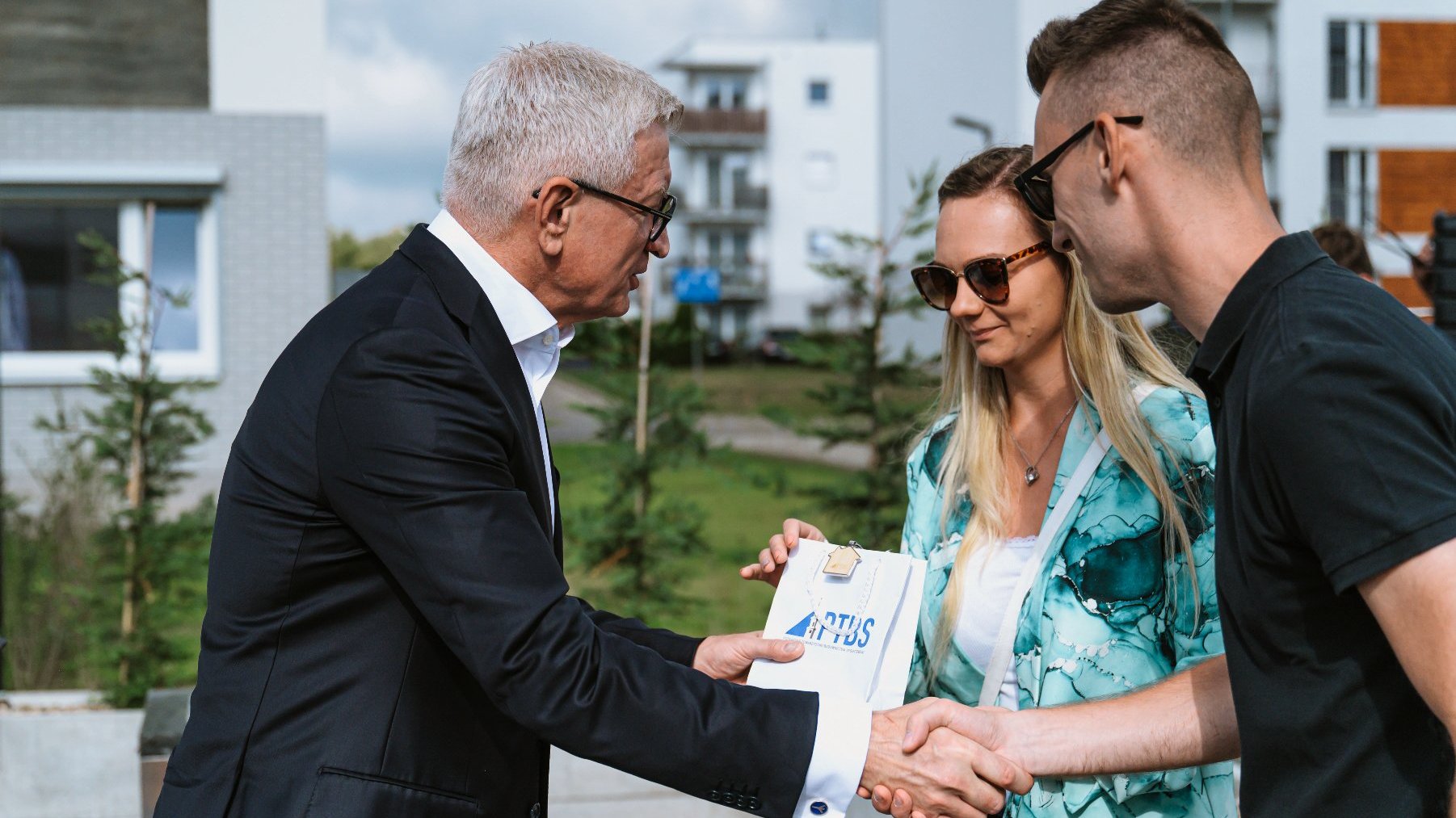 Na zdjęciu widać kobietę i mężczyznę, którzy odbierają klucze od Prezydenta Poznania. Mężczyzna i Prezydent podają sobie rękę. W tle widać bloki mieszkalne.