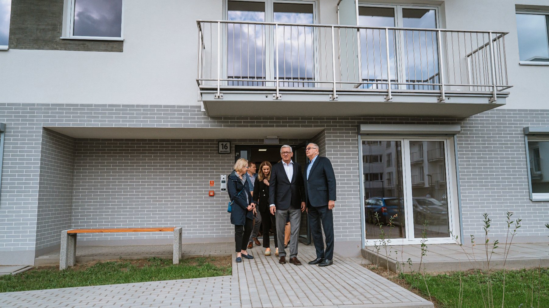 Na zdjęciu widać grupę osób, która wychodzi z budynku na dwór. Budynek ma numer 16. Na drzwiami wejściowymi do budynku jest balkon. Obok drzwi wejściowych jest ławka.