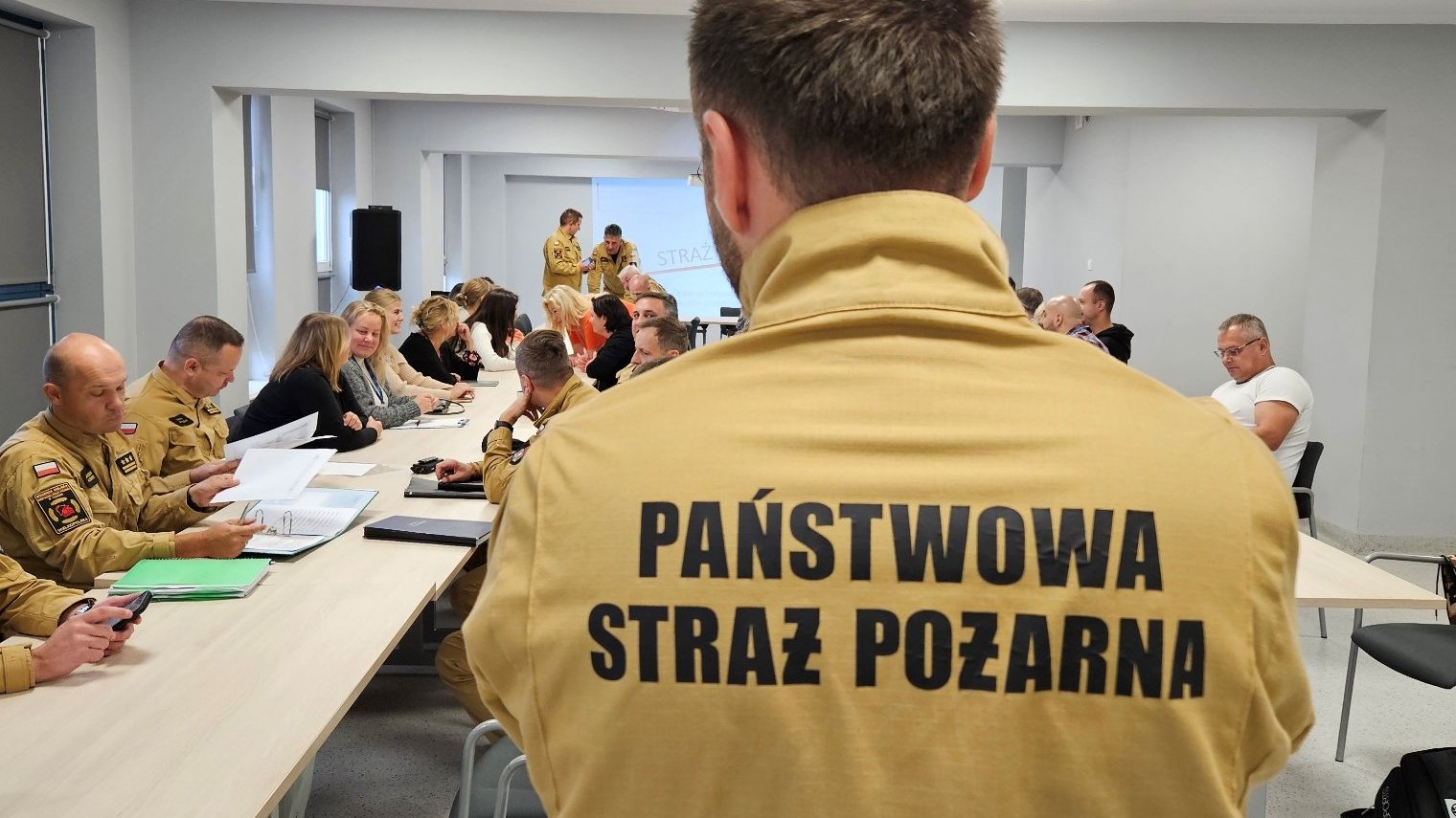 Na zdjęciu widać ludzi podczas szkolenia w sali. Na pierwszym planie jest osoba w żółtej koszulce z napisem Państwowa Straż Pożarna. Osoba ta stoi tyłem do obiektywu. W tle zdjęcia widać inne osoby, które siedzą przy stołach.