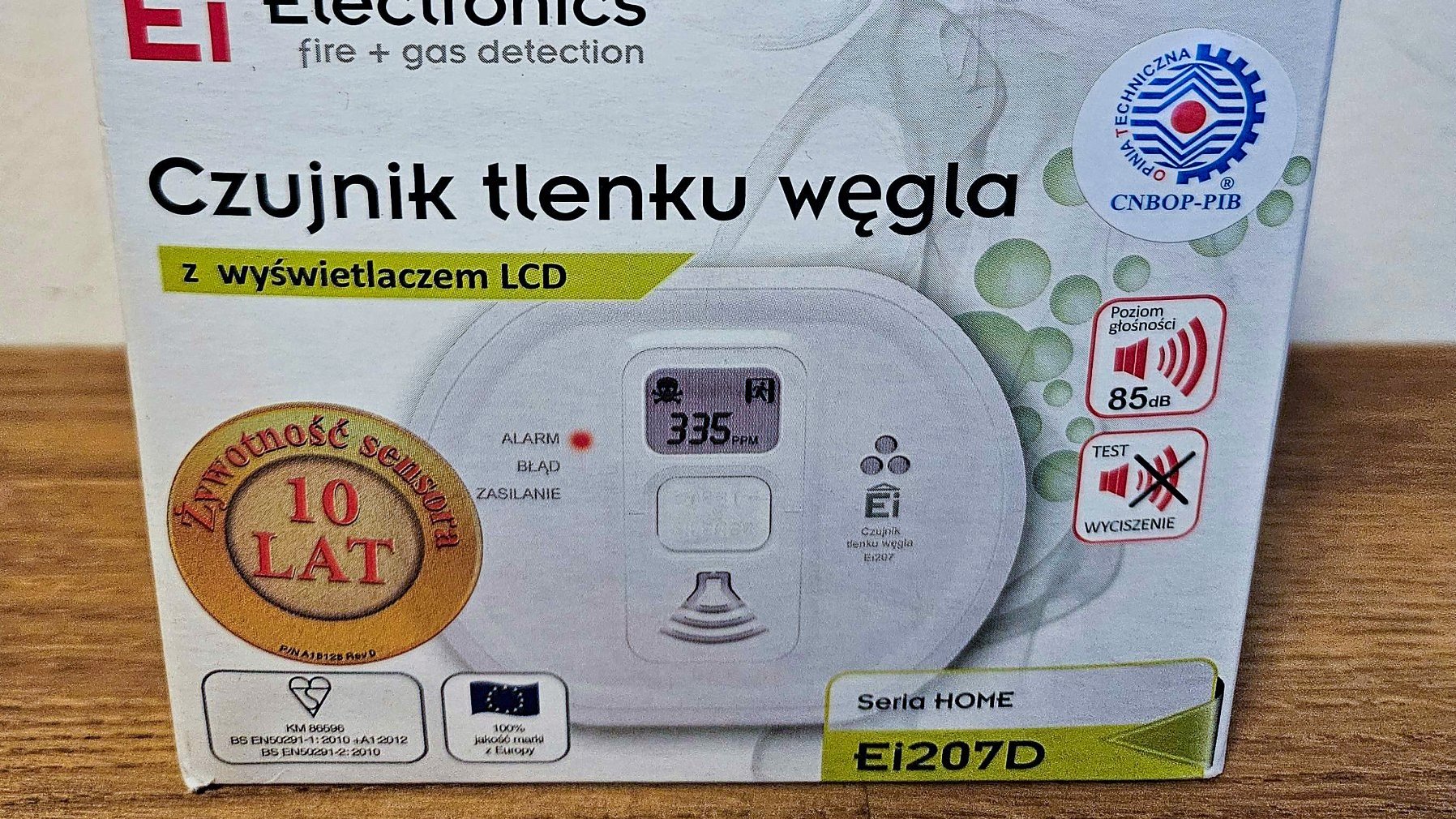 Na zdjęciu widać przód kartonowego opakowania do czujnika czadu. Opakowanie leży na drewnianym stole. Na opakowaniu znajduje się informacja o marce czujnika, opis parametrów czujnika i zdjęcie czujnika.