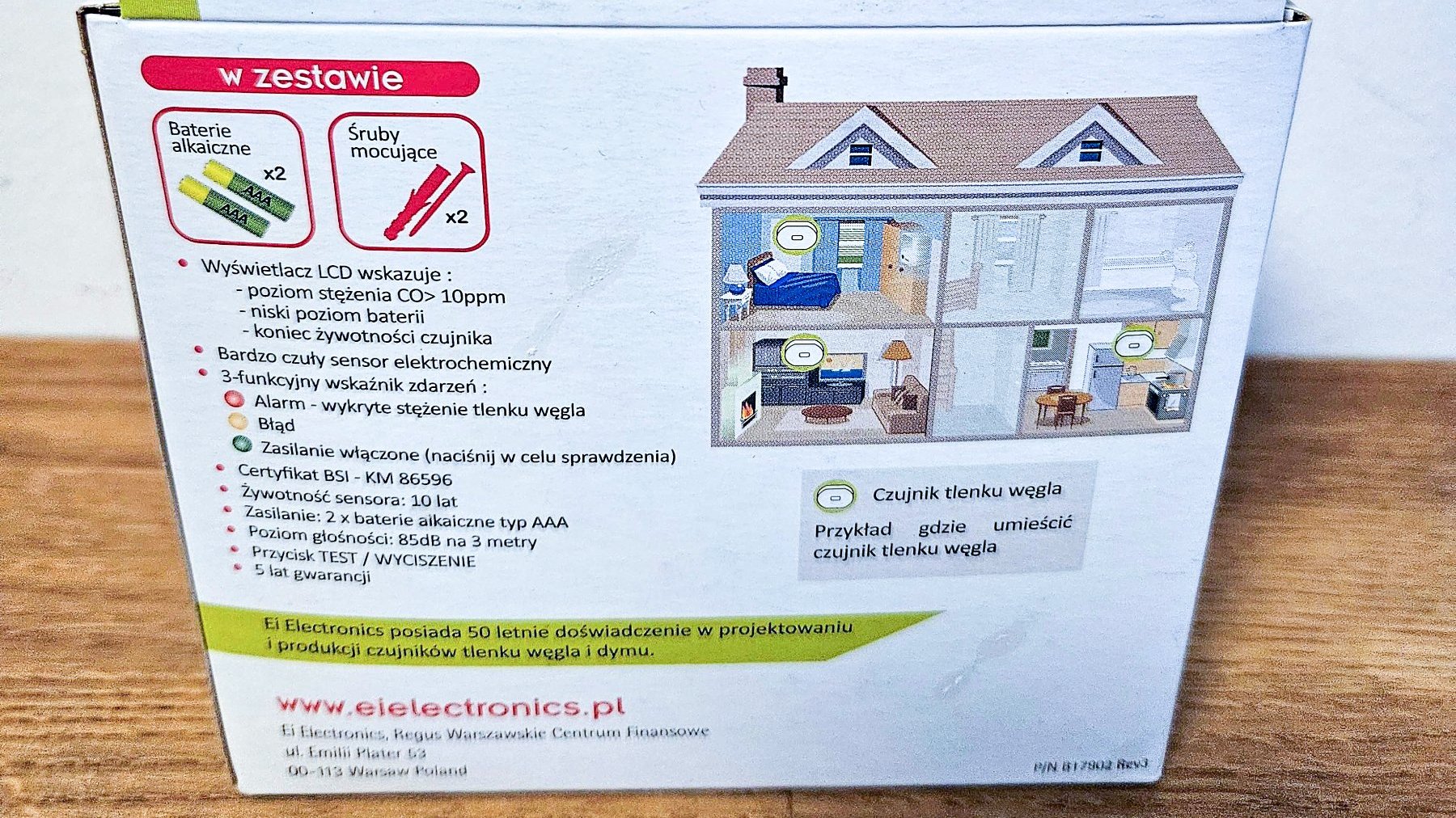 Na zdjęciu widać tył kartonowego opakowania do czujnika czadu. Opakowanie leży na drewnianym stole. Na opakowaniu znajduje się informacja o elementach, które są w zestawie z czujnikiem, szczegółowy opis parametrów czujnika i rysunek domu z zaznaczonymi miejscami, gdzie można zainstalować czujnik.
