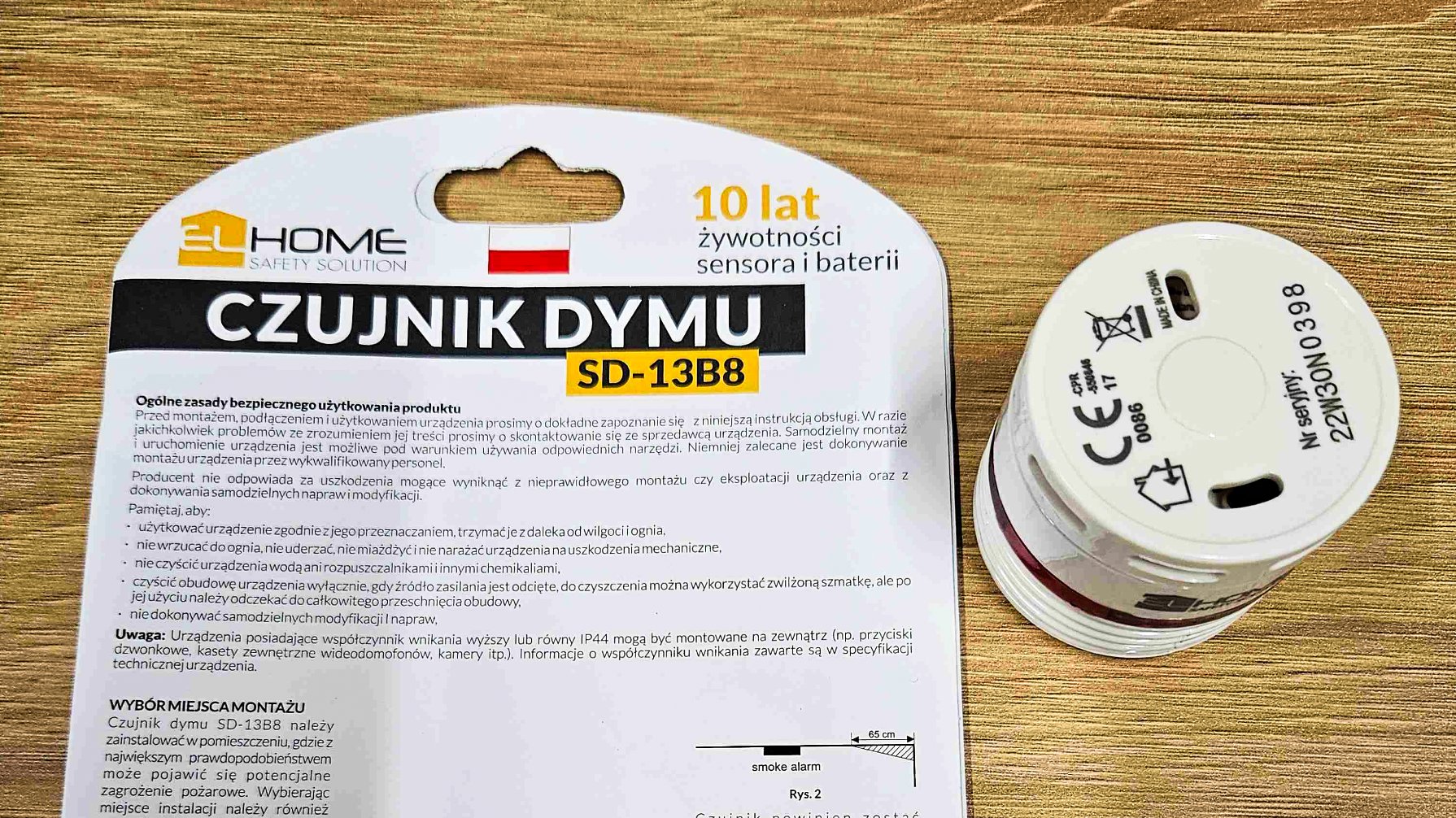 Na zdjęciu widać czujnik dymu i instrukcję jego używania. Czujnik jest w formie walca, w białym kolorze. Na instrukcji wypisane są zasady bezpiecznego używania czujki. Obie rzeczy leżą na drewnianym stole.
