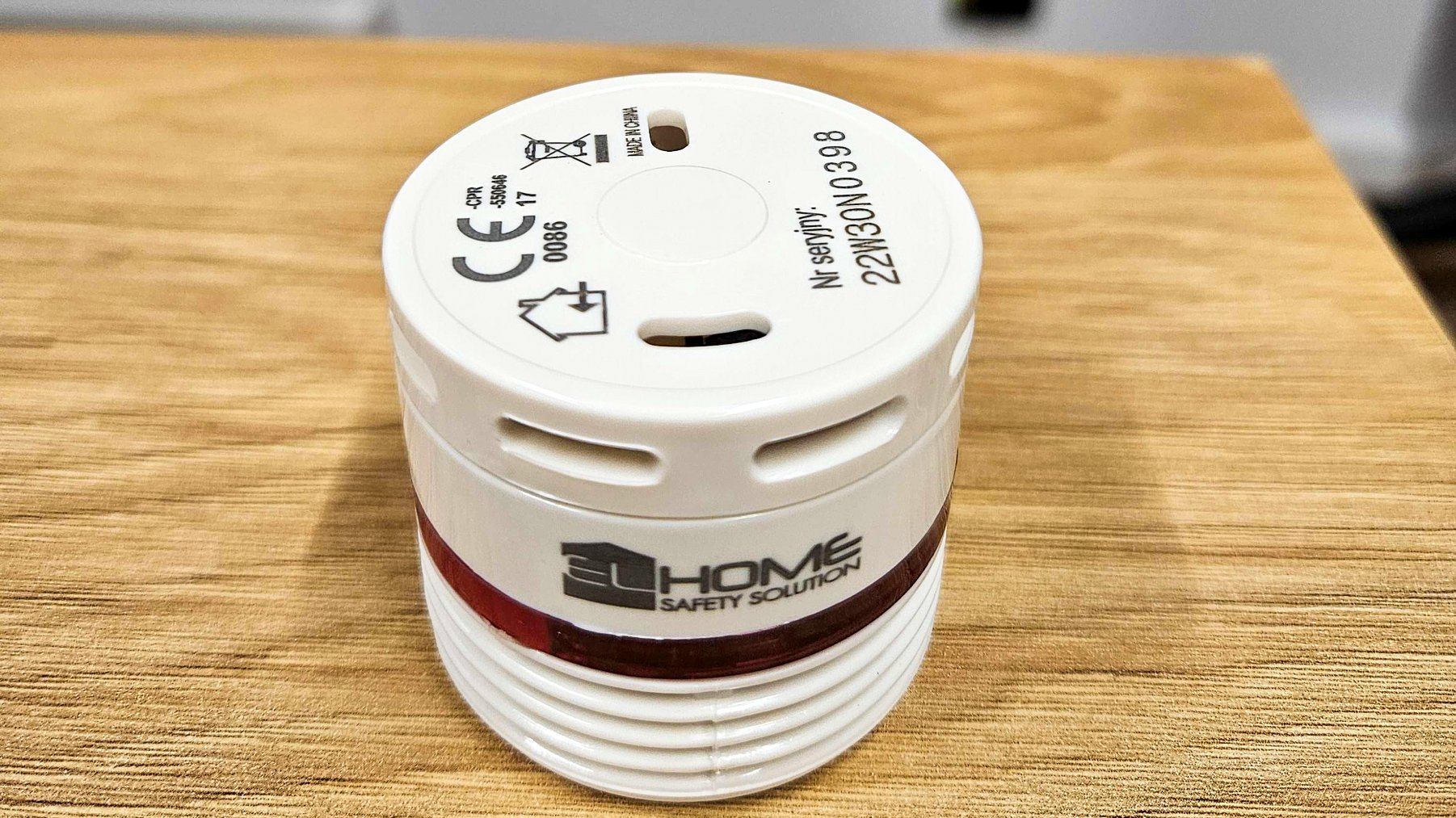 Na zdjęciu widać czujkę dymu. Czujka jest w kształcie walca, w białym kolorze, z napisem Home Safety Solution. Czujka stoi na drewnianym stole.