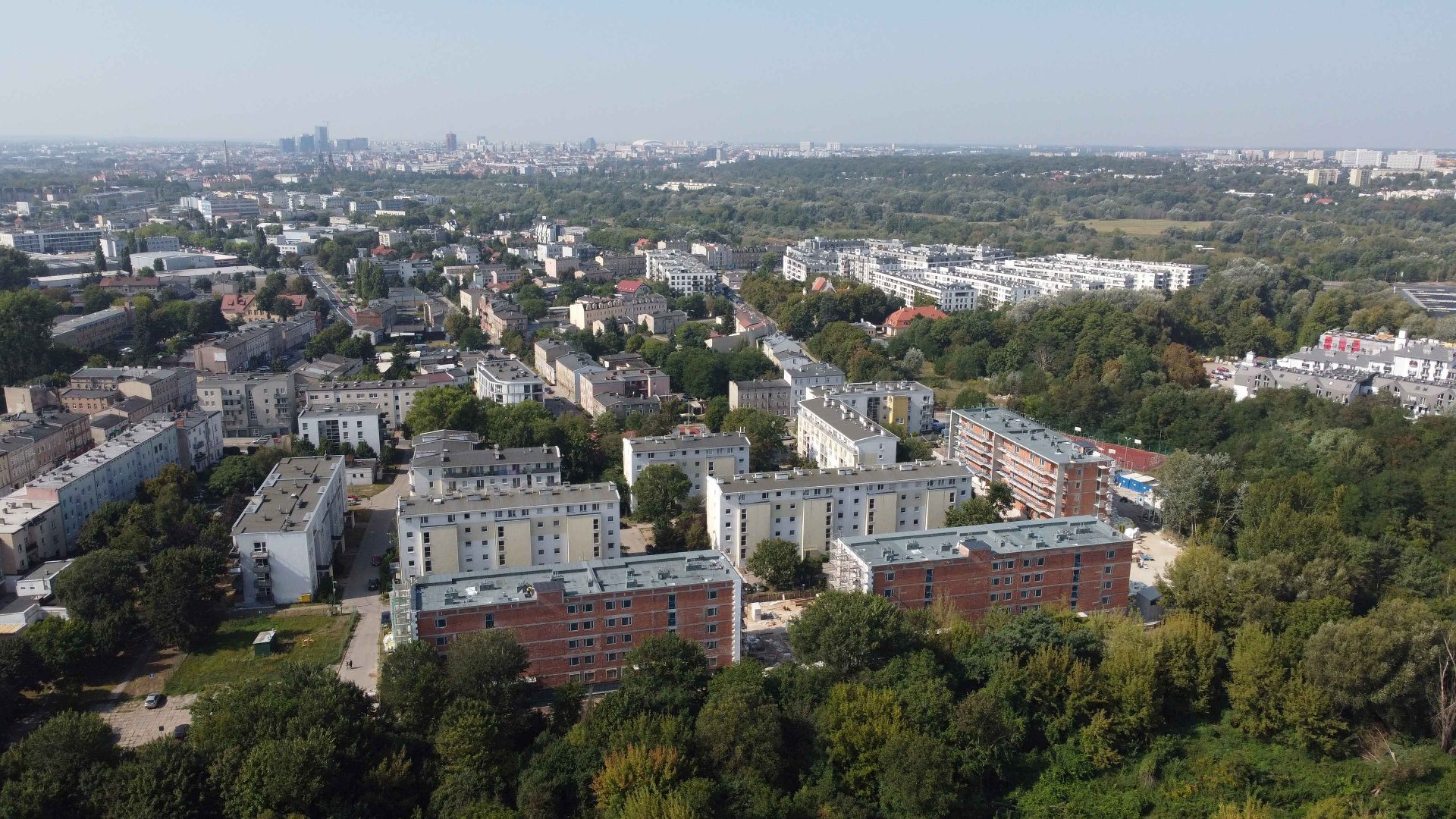 Na zdjęciu widać panoramę Poznania. Z przodu i z prawej strony widać duży obszar zieleni. Na środku i z lewej strony widać budynki mieszkalne. 3 budynki są w trakcie budowy. Reszta budynków jest wykończona.