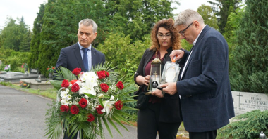 Od lewej: mężczyzna w garniturze trzymający bukiet kwiatów, kobieta w okularach trzymająca jedną znicz i mężczyzna zapalający drugą. Cała trójka znajduje się na ścieżce cmentarnej przed grobem poległego podczas Czerwca '56.