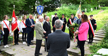Na zdjęciu grupa ludzi stojąca przed grobami, są sztandary, kwiaty oraz logo szkoły