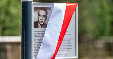 Tabliczka z informacją przyczepiona do słupa w parku przepasana wstęgą biało-czerwoną.