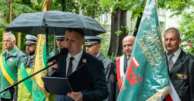 Na zdjęciu dyrektor Gabinetu Prezydenta przy mikrofonie, w tle ludzie z parasolami