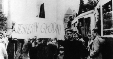 Czarno-biała fotografia strajkujących robotników. Wspólnie trzymają transparent z hasłem "jesteśmy głodni".