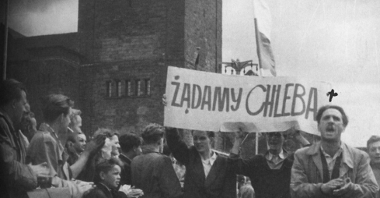 Czarno-biała fotografia strajkujących robotników. Wspólnie trzymają transparent z hasłem "Żądamy chleba". Uczestnicy stoją pod Zamkiem Cesarskim.