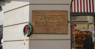 Miedziana tablica powieszona na rogu budynku. Tablica jest prostokątną płytą, z napisem: "Romek Strzałkowski 1943-1956. Chłopiec bohater. Zginął od kuli w dniach robotniczego protestu".