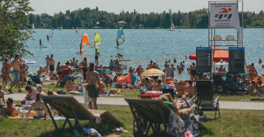 Jezioro Rusałka. Tłum osób siedzi na plaży nad Rusałką. W tle widok na jezioro. Po jeziorze pływają żaglówki.