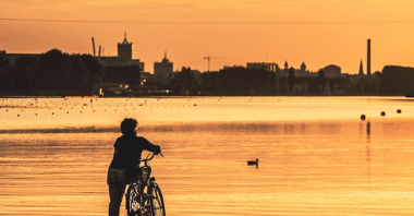 Zachód słońca nad Jeziorem Maltańskim. Na pierwszym planie kobieta z rowerem, stoi plecami. W tle Jezioro Malta oraz panorama miasta.