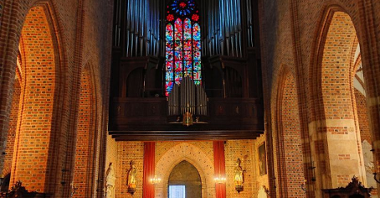 Organy w Katedrze Poznańskiej. Znajdują się nad głównym wejściem do katedry. Za organami wysoki, kolorowy witraż. Pod spodem stoją ławki.