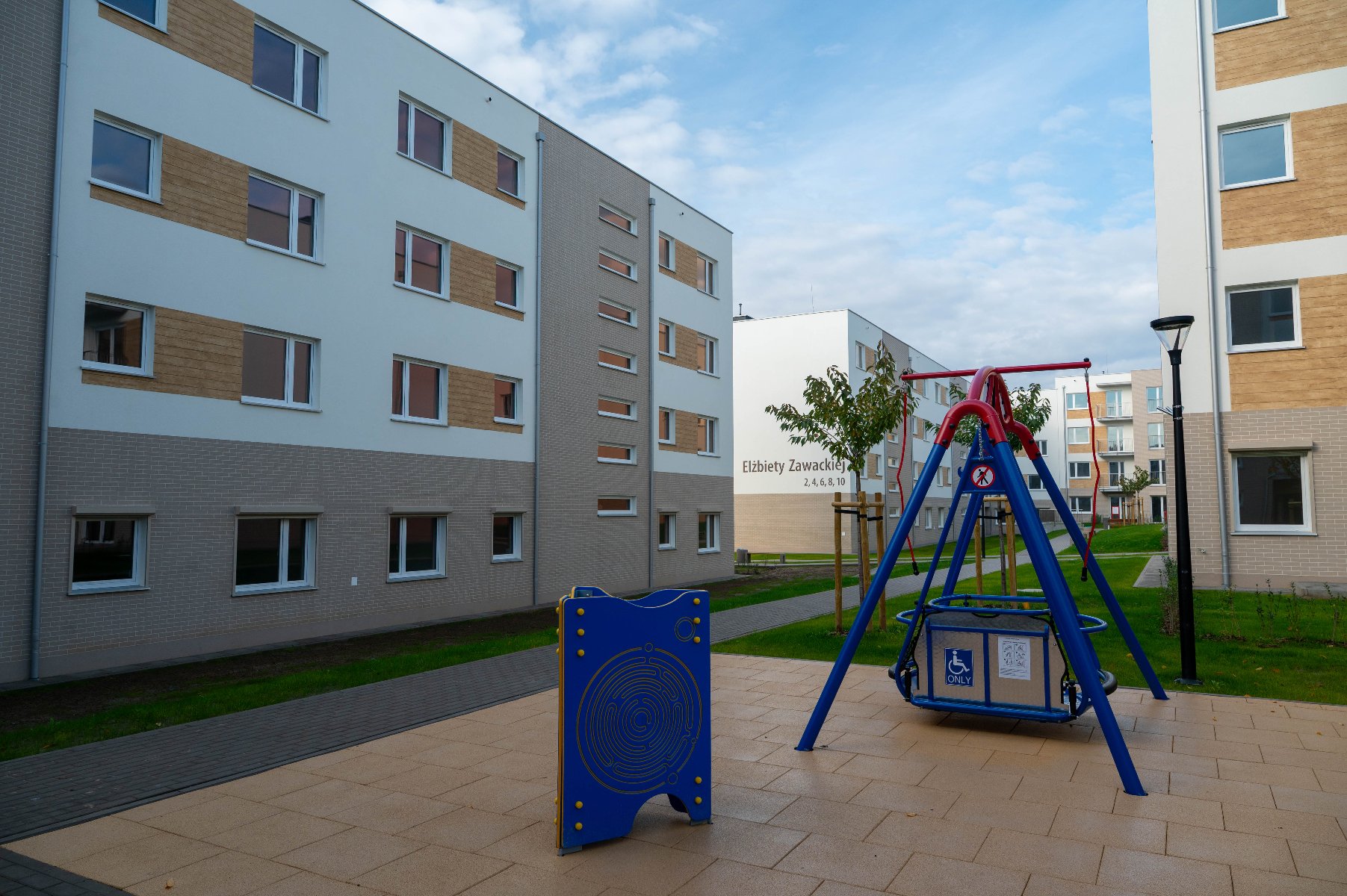 Zakończyła się budowa II etapu inwestycji mieszkaniowej realizowanej w ramach miejskiego programu "POZnań - i zamieszkaj" Plac zabaw z czerwoną i niebieską huśtawką przystosowaną dla osób z niepełnosprawnościami na tle nowoczesnych, jasnych budynków mieszkalnych. Na jednym z budynków widoczny napis: "Elżbiety Zawackiej". Po lewej stronie niebieski panel edukacyjny.