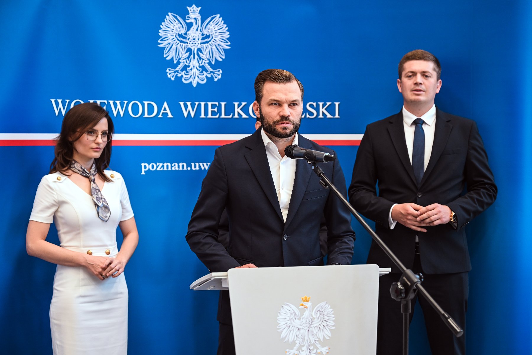 Miasto Poznań otrzyma kolejne 219 milionów złotych bezzwrotnego dofinansowania na rozwój budownictwa komunalnego rzy osoby w formalnych strojach stoją na tle niebieskiego baneru z napisem "WOJEWODA WIELKOPOLSKI". Mężczyzna w centrum przemawia do mikrofonu, a kobieta w białej sukience stoi po jego lewej stronie. Na mównicy znajduje się godło.