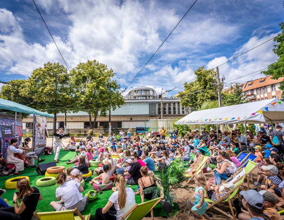 Lato na Madalinie świętuje już 10 lat! Organizatorzy przygotowali coś specjalnego na rozpoczęcie tegorocznych wakacji. Tłum dzieci i dorosłych siedzi na plażowych leżakach przez sceną. Na scenie dwuosobowy zespół, kobieta gra na wiolonczeli a mężczyzna na gitarze. Za nimi dekoracje. W tle budynki i wysokie, zielone drzewa. Niebo jest blękitne i pokryte białymi chmurami.