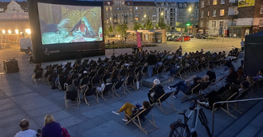 Wieczór na miejskim rynku, ludzie siedzą na krzesełkach i oglądają film na ekranie wielkoformatowym