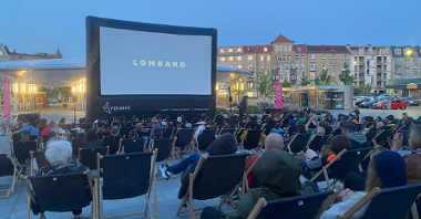 Wieczór na miejskim rynku, ludzie siedzą na krzesełkach i czekają na pokaz filmu Lombard na ekranie wielkoformatowym