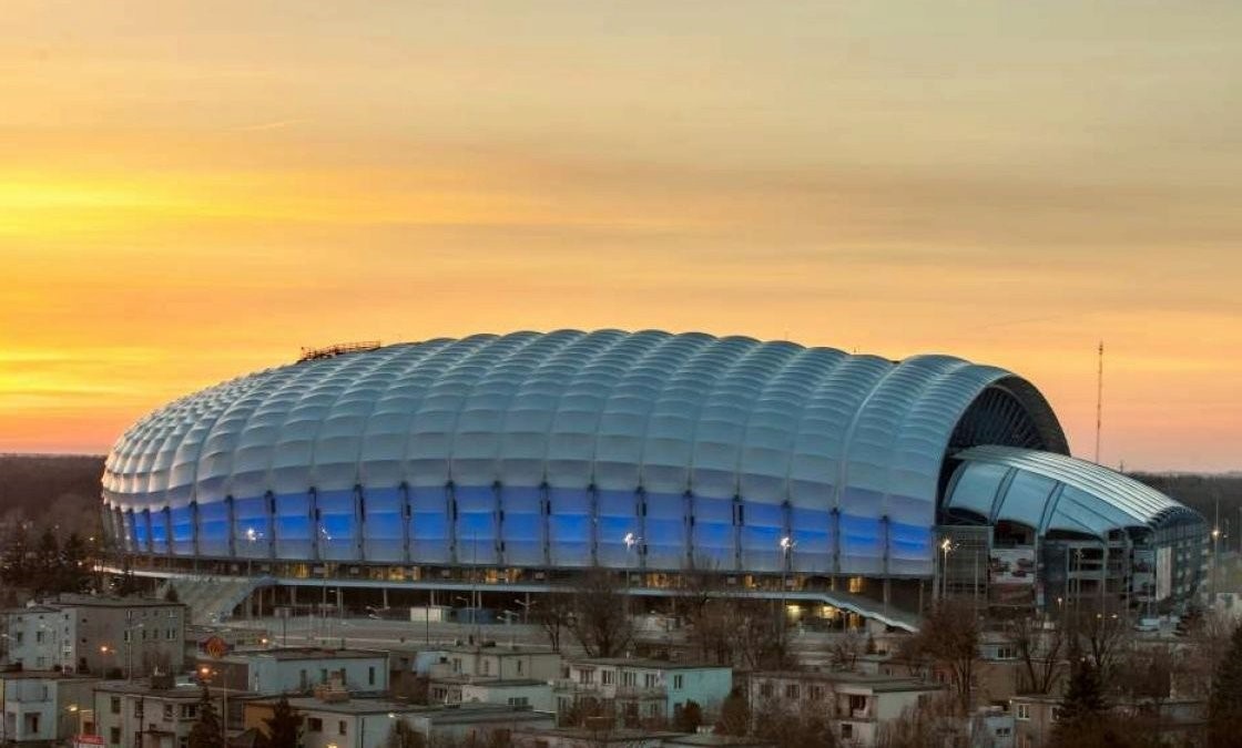 widok bryły stadionu przy ul. Bułgarskiej w Poznaiu, w otoczeniu budynków mieszkalnych, na tle nieba rozświetlonego przez zachodzące słońce - grafika artykułu