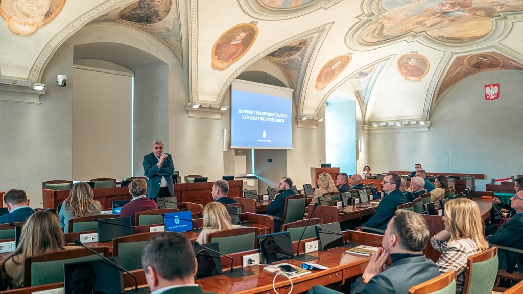 w sali sesyjnej przy stanowiskach z monitoriami i mikrofonami siedzą osoby, przed nimi stoi jedna osoba - mężczyzna trzymający mikrofon, sklepienie sali zdobią kolorowe, zabytkowe freski