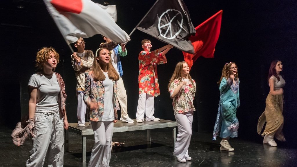 spektakl teatralny z udziałem młodych ludzi, niektórzy z nich trzymają fagi, na jednej z nich symbol pacyfizmu