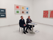 dwóch mężczyzn siedzi przy stoliku w białej sali, na jej ścianach wiszą obrazy przedstawiające sztukę nowoczesną