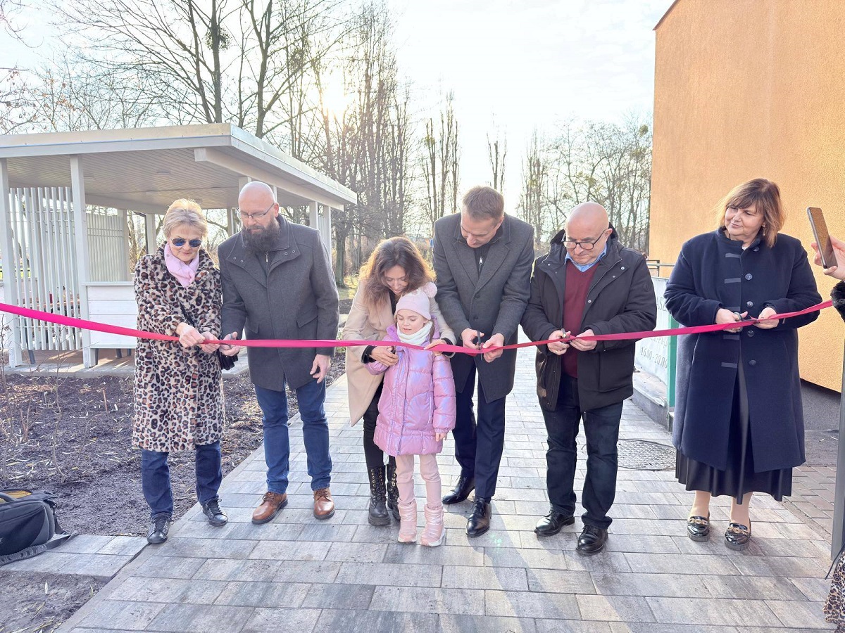 grupa osób kobiet, mężczyzn, dziecko przecinaja wstęgę na terenie przed budynkiem przedszkola