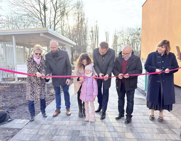 grupa osób kobiet, mężczyzn, dziecko przecinaja wstęgę na terenie przed budynkiem przedszkola