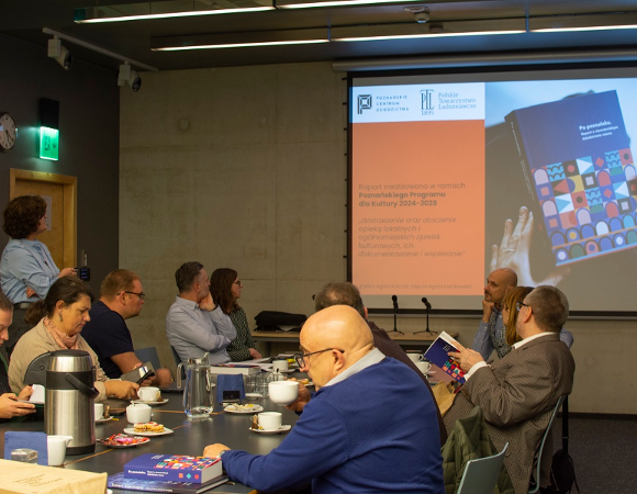 grupa osób w sali konferencyjnej siedzi przy stole i ogląda prezentację multimedialną na ekranie