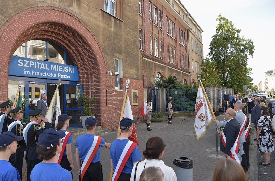 Uroczystość przed szpitalem im. F. Raszei grupa osób ze sztandarami i szarfami w biało czerwonych kolorach stoi przed wejściem do budynku szpitala