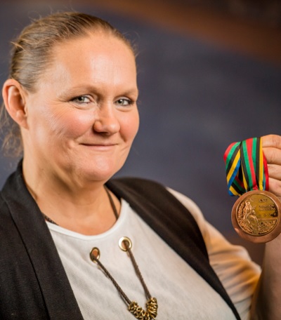 Na zdjęciu kobieta, włosy spiętedo tyłu, uśmiecha się, w ręku trzyma medal na kolorowej szarfie