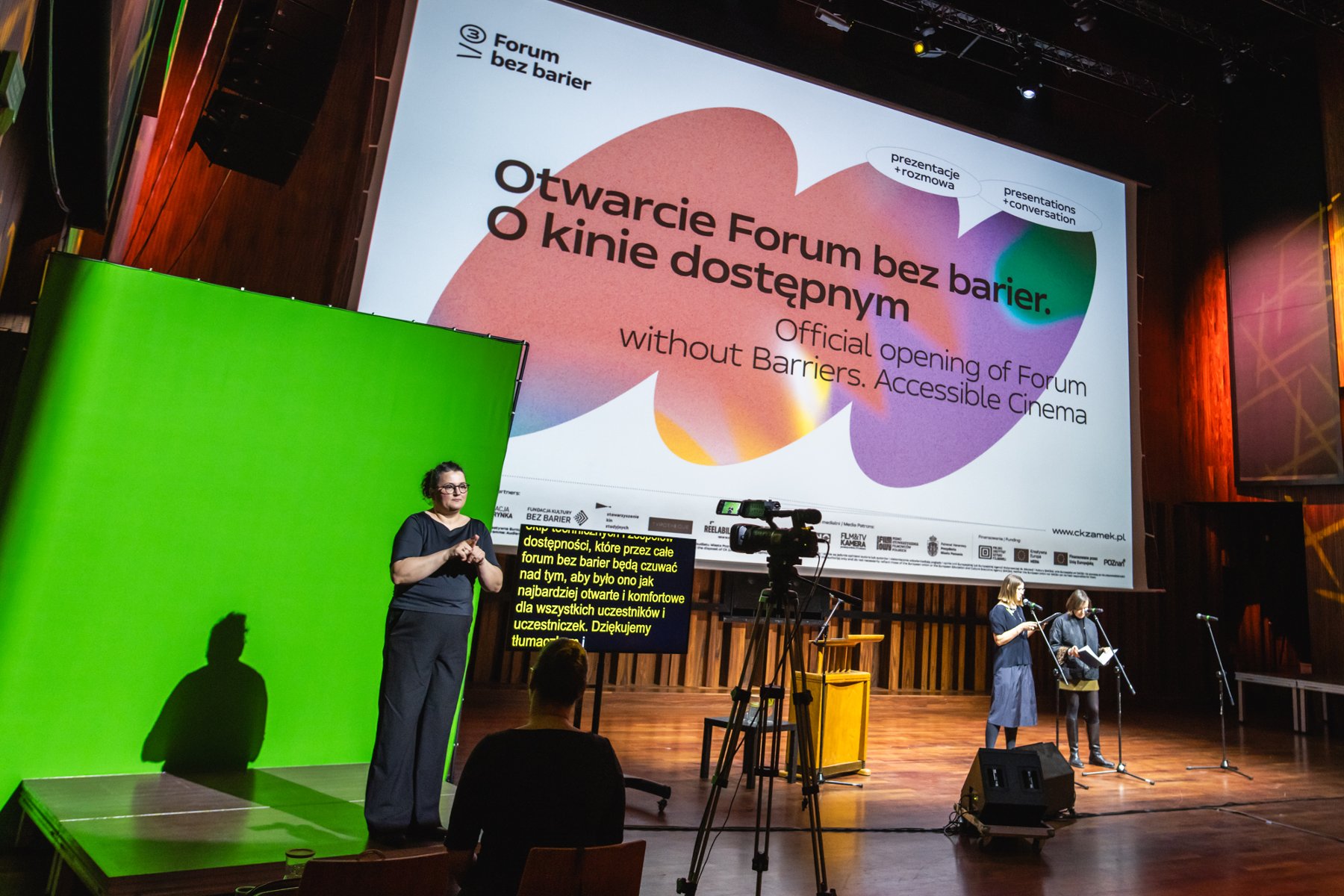 Na zdjęciu tłumacz polskiego języka migowego na zielonym tle. W tle plansza z informacją o otwarciu forum.