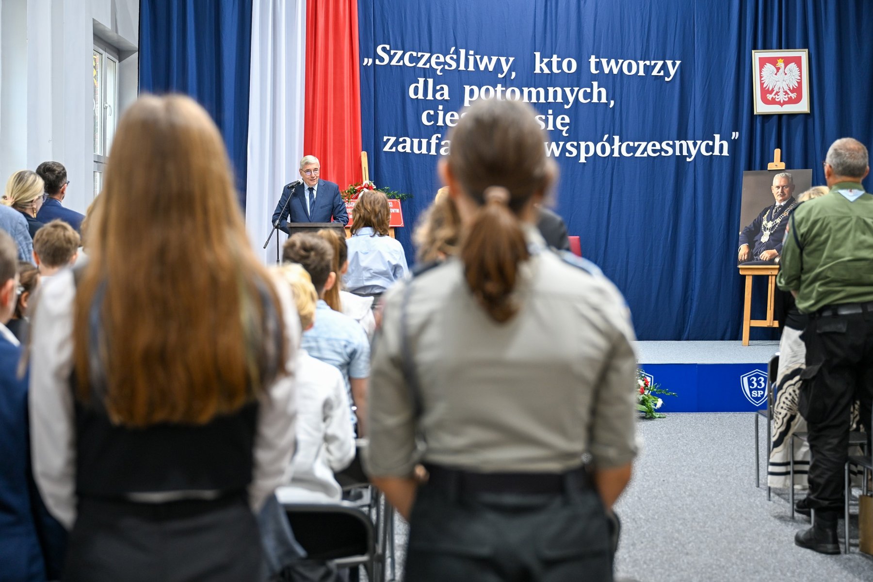 Zdjęcie przedstawia przemawiającego podczas szkolnej akademii przewodniczącego rady miasta. - grafika artykułu