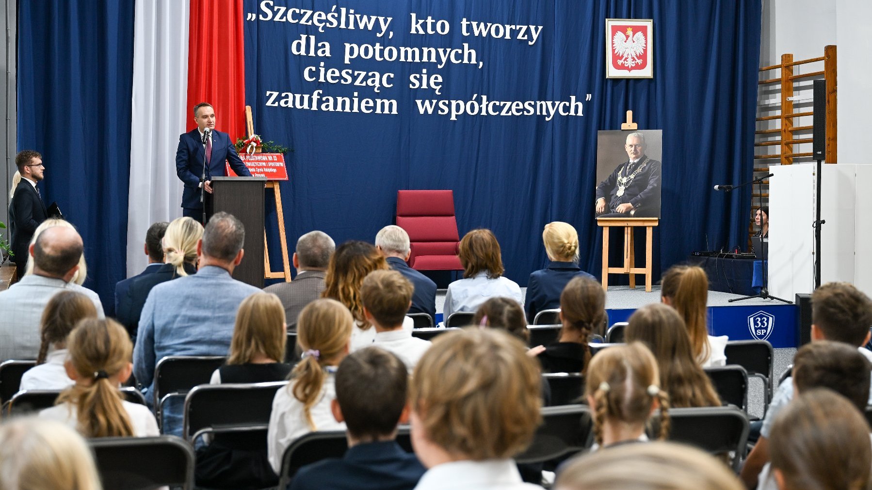 Zdjęcie przedstawia przemawiającego podczas szkolnej akademii zastępcę prezydenta miasta.